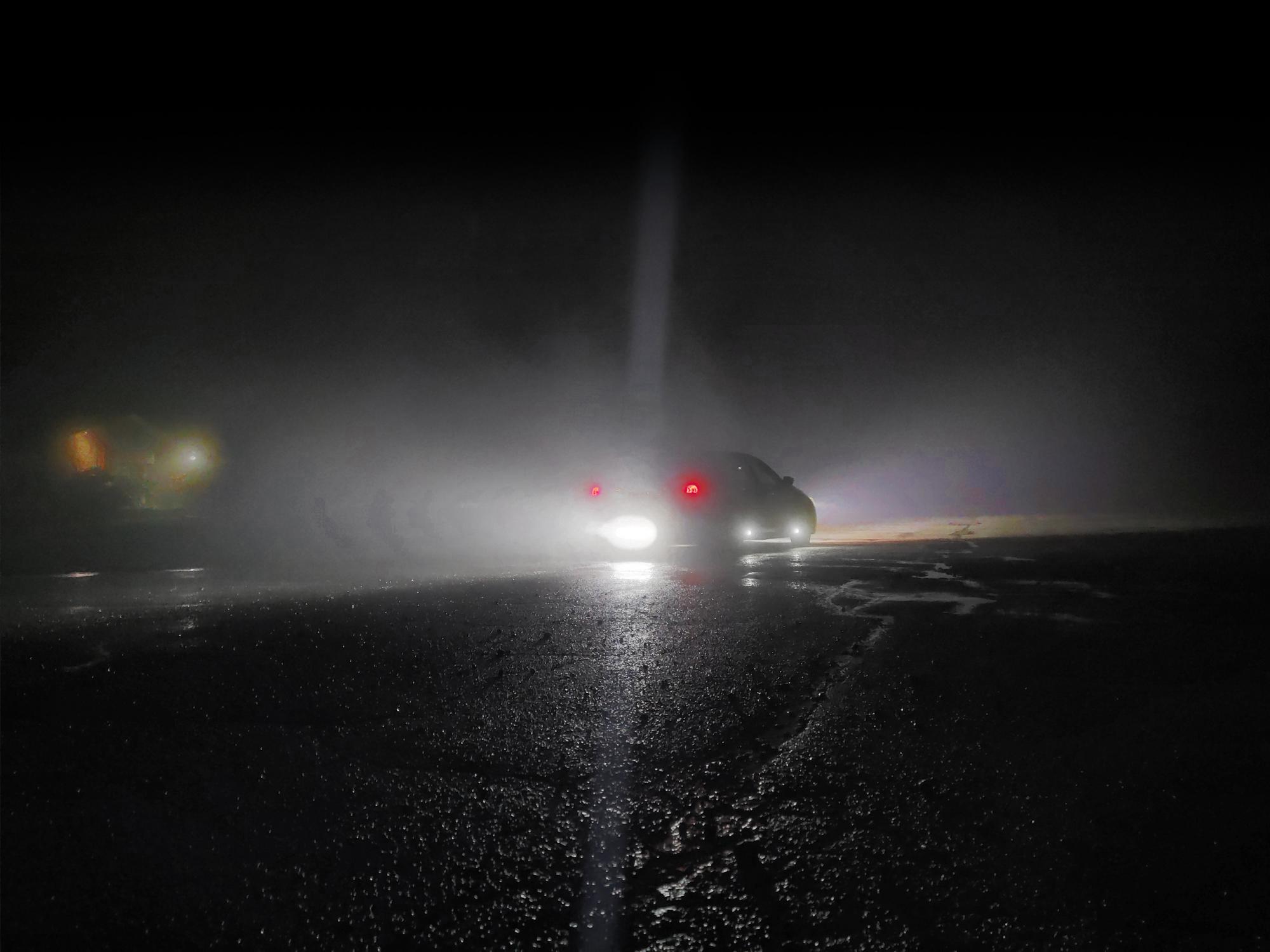A sedan in a dark parking lot pictured at a distance from the back right corner, with blinding lights on the rear, sides, and front of the car flooding the surrounding area with cool white light.