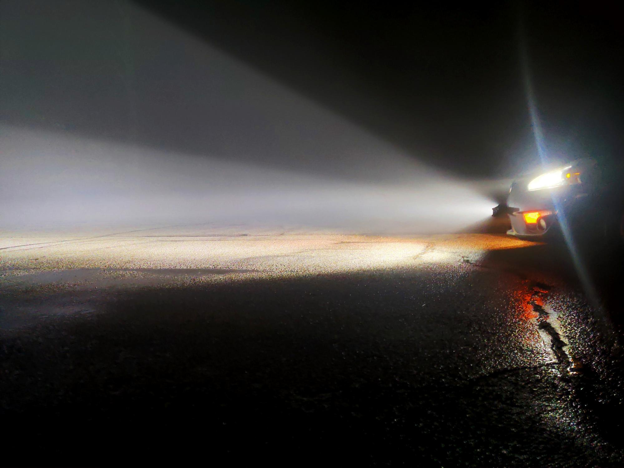 A side profile of the front of a sedan in a dark foggy parking lot, with bright lightbar casting a cone of light ahead.