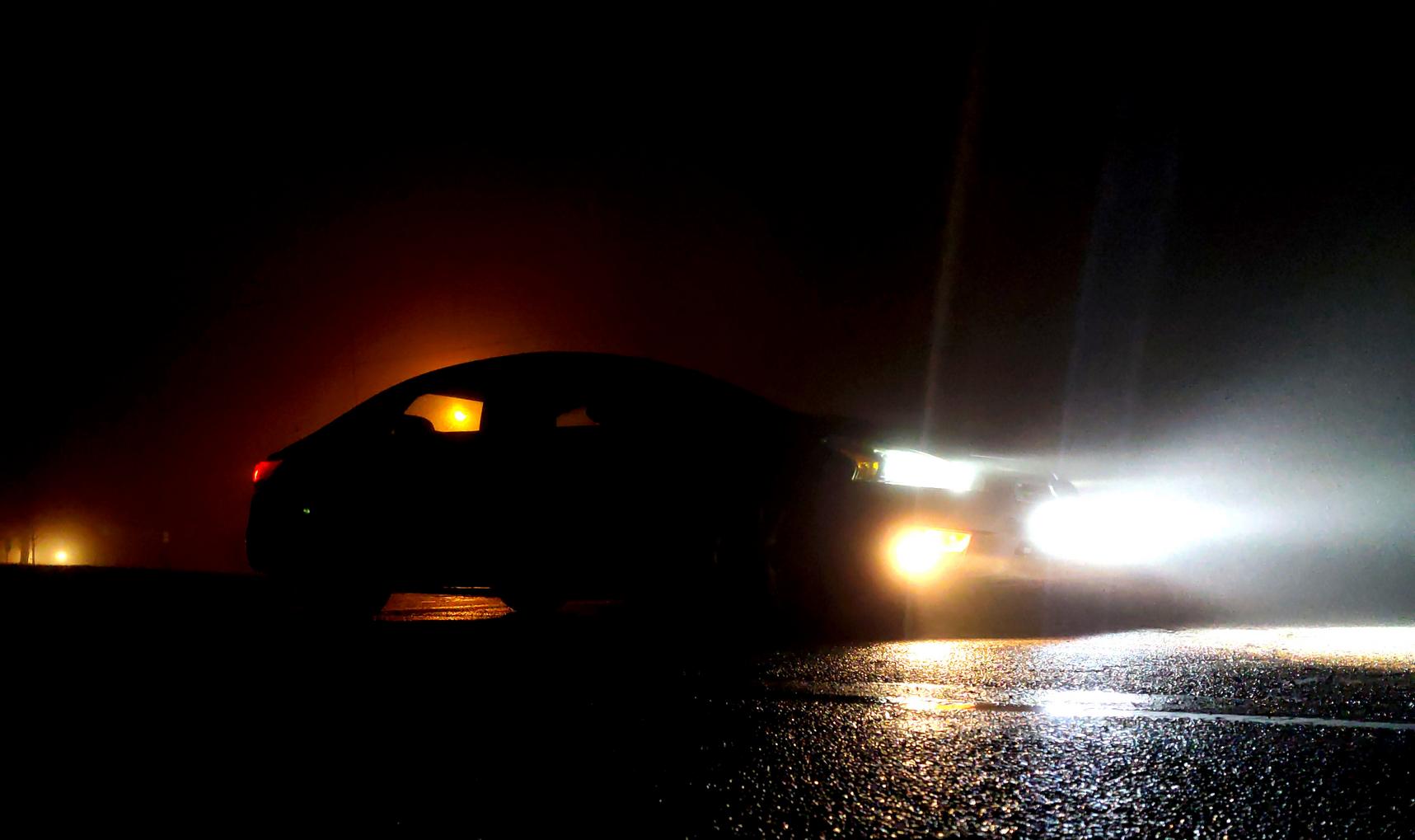 A street-lamp back-lit sedan in a dark foggy parking lot, pictured from the front right corner, with head lights, fog lights, and a center light bar illuminating the area ahead.