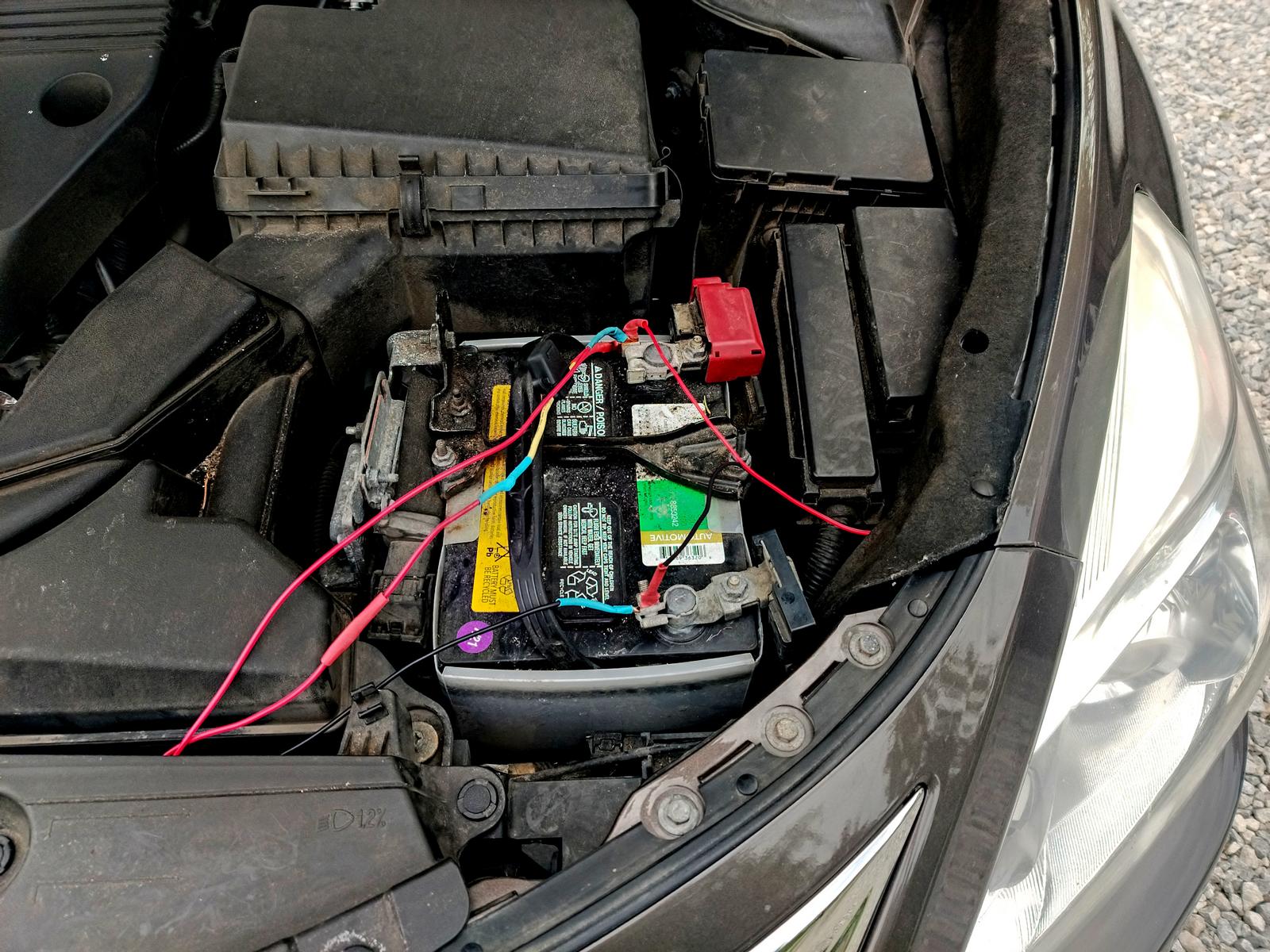 A picture of a car's engine bay, focused on the 12 volt battery.